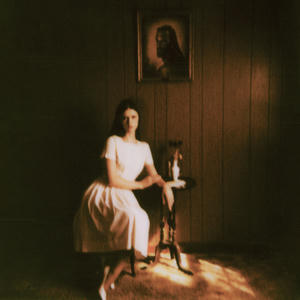 Cover art of Ethel Cain's Preacher's Daughter.
It depicts Ethel sitting on a chair wearing a white dress.
Her arm is resting on a side table.
Above her on the wall hangs a portrailt of a man with a beard and long hair.