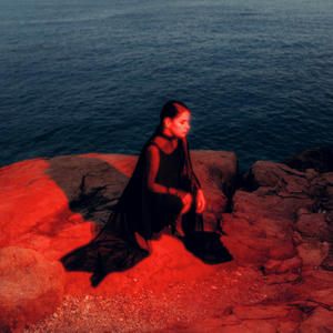 Cover art of Sofia Kourtesis' Madres.
It shows Sofia sitting by a cliff. The sea can be seen in the background.