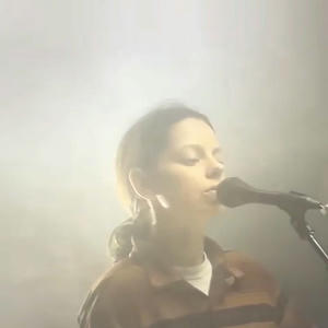 Cover art of Tirzah's trip9love...???.
It depicts a woman standing in front of a microphone.
The background is foggy and unclear, as if taken during a concert with a lot of smoke machines.
The woman has her hair in a ponytail and is wearing a brown flannel on top of a white shirt.