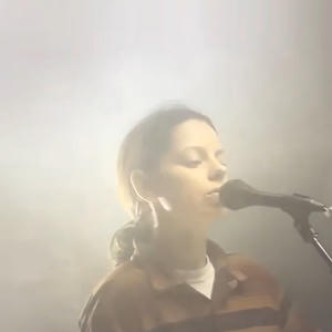 Tirzah - trip9love...??? Cover art of Tirzah's trip9love...???.
It depicts a woman standing in front of a microphone.
The background is foggy and unclear, as if taken during a concert with a lot of smoke machines.
The woman has her hair in a ponytail and is wearing a brown flannel on top of a white shirt.